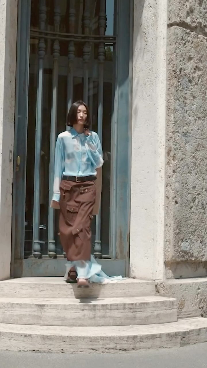 A woman in a sheer blue blouse and brown skirt stands on stone steps outside an iron gate in bright daylight.