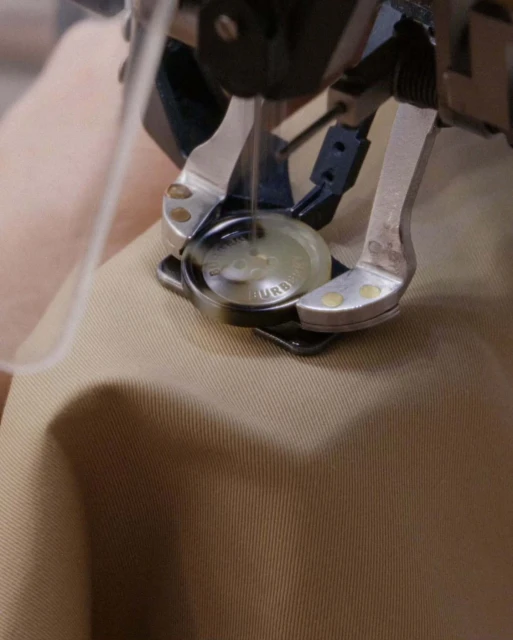 Close-up of a sewing machine applying a button to tan fabric. The needle and pressure foot are visible, highlighting detail and texture.