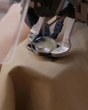 Close-up of a sewing machine applying a button to tan fabric. The needle and pressure foot are visible, highlighting detail and texture.
