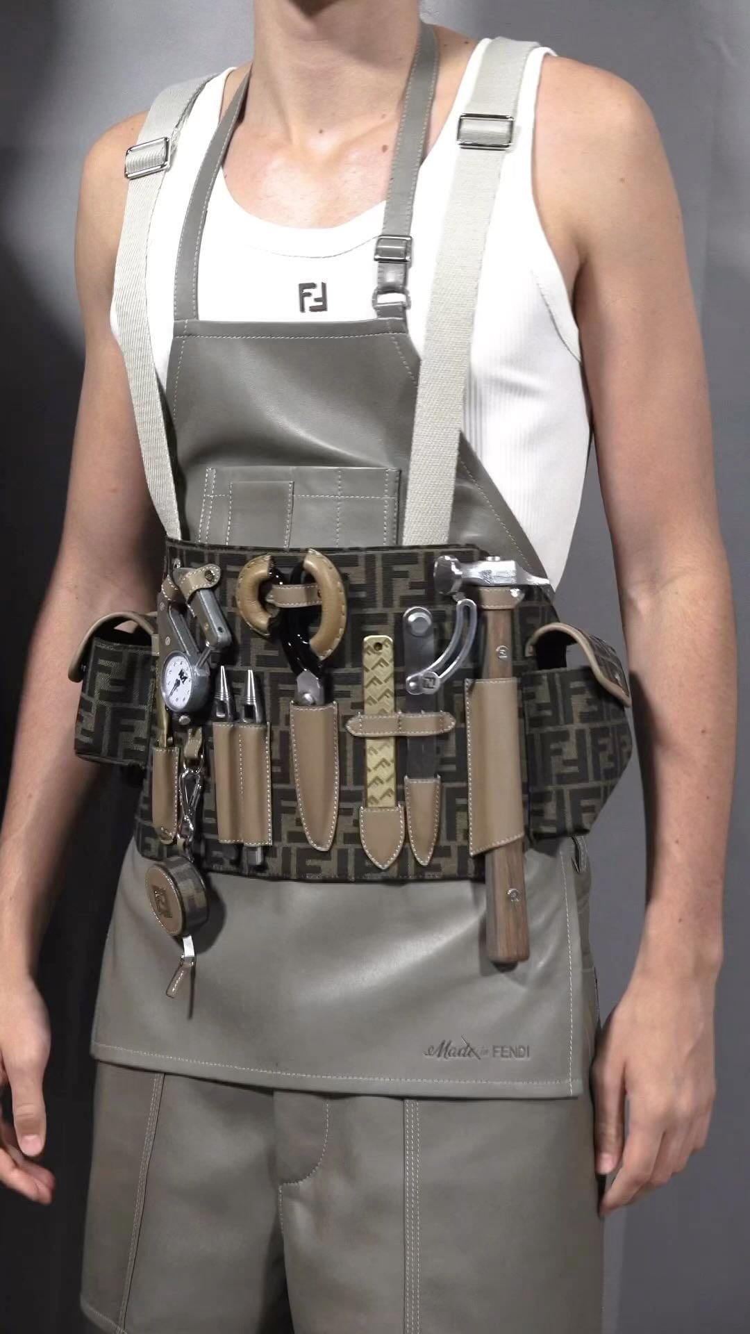 Man wearing a Fendi logo utility apron and shorts, accessorized with tools, standing in a studio against a grey backdrop.