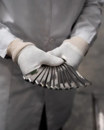 Person in lab coat and gloves holding a fan of metal cutlery, displaying tableware in a neutral indoor setting.