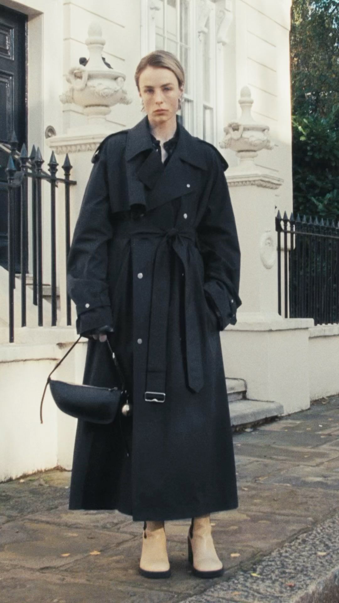 A woman wearing a long black trench coat and light-colored heeled boots stands outside a building with black iron gates.