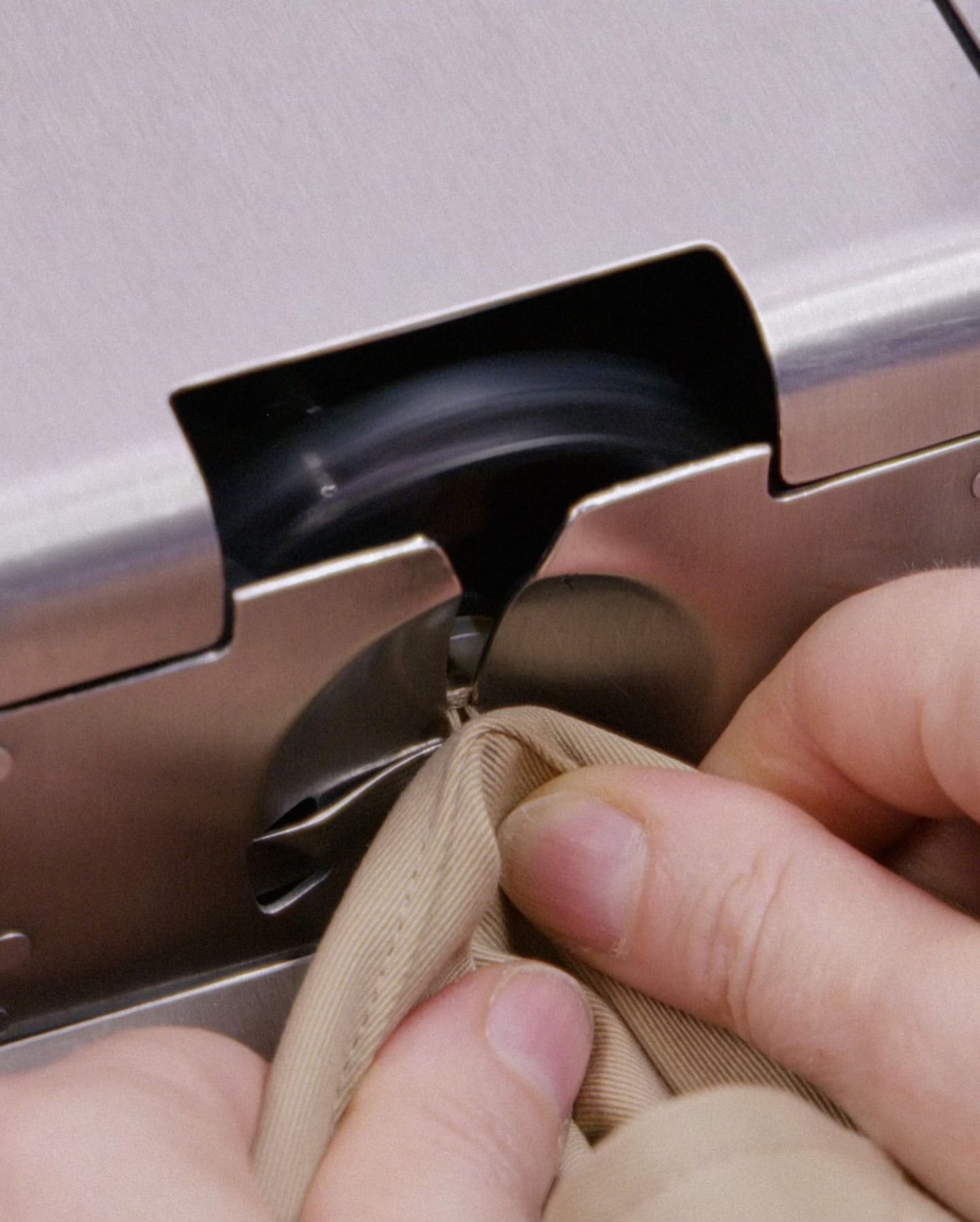 Close-up of a person using a metal machine to attach a metal snap to a piece of tan fabric, focusing on the device and the hands guiding the fabric.