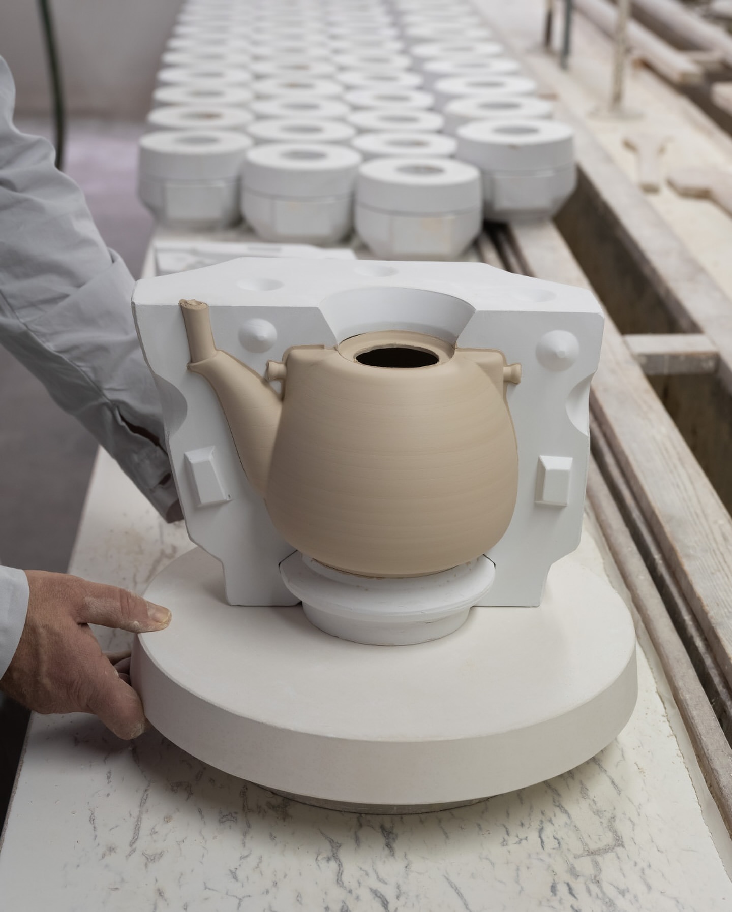 Craftsperson creating a ceramic teapot in a plaster mold, production line of molds in background, neutral color palette.