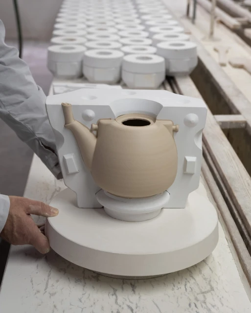Craftsperson creating a ceramic teapot in a plaster mold, production line of molds in background, neutral color palette.