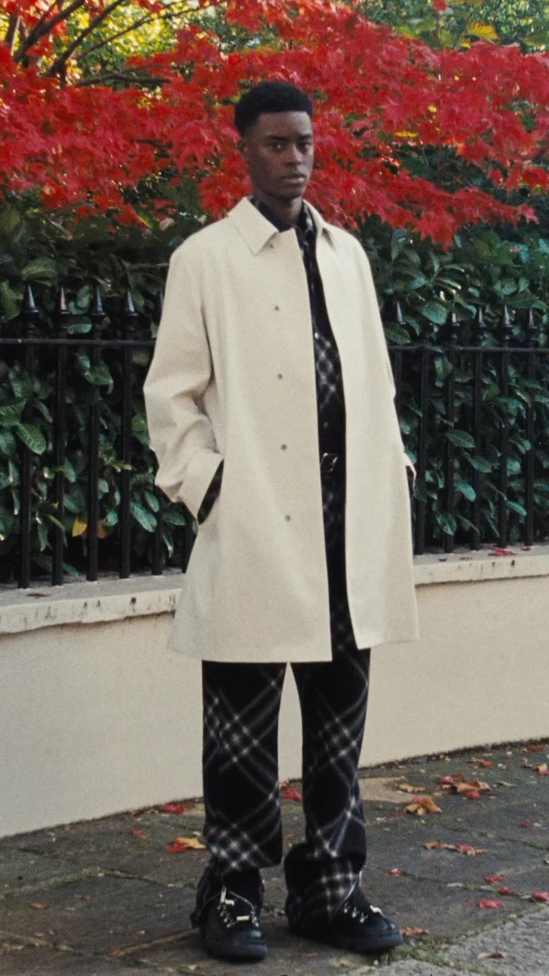 A Black man standing outdoors wearing a cream coat and dark plaid trousers in front of a low wall and autumn foliage.