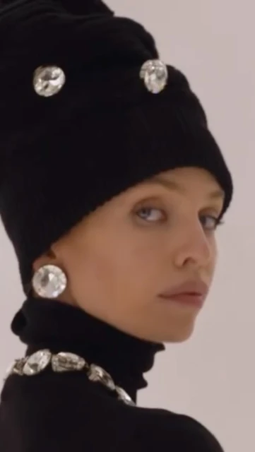 Headshot of a fashionable woman wearing a black turtleneck, jeweled necklace, earrings, and hat with matching jewels, against a bright background.
