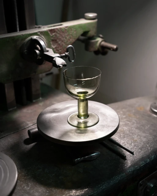 Close-up of a clear glass goblet sitting on a shiny metal turntable of a vintage glass-cutting lathe in a dimly lit workshop or studio.