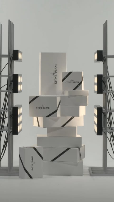 A stack of Stone Island branded boxes is displayed against a neutral background, flanked by industrial lighting fixtures and wiring.