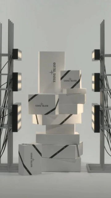 A stack of Stone Island branded boxes is displayed against a neutral background, flanked by industrial lighting fixtures and wiring.
