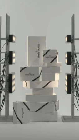 A stack of Stone Island branded boxes is displayed against a neutral background, flanked by industrial lighting fixtures and wiring.