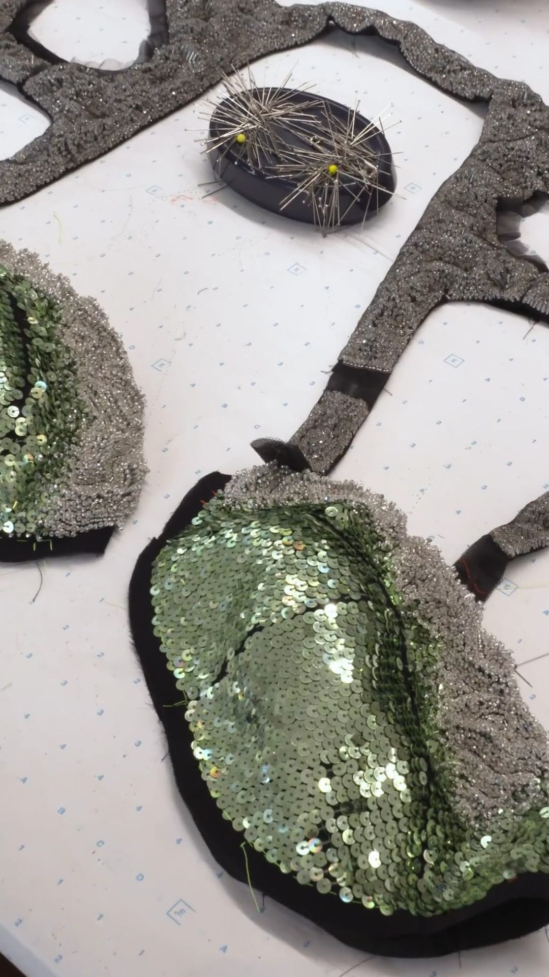 Close-up of fabric pieces with black trim, embellished with shiny green sequins and silver-tone beads, along with a black pin cushion.