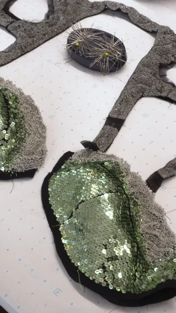 Close-up of fabric pieces with black trim, embellished with shiny green sequins and silver-tone beads, along with a black pin cushion.