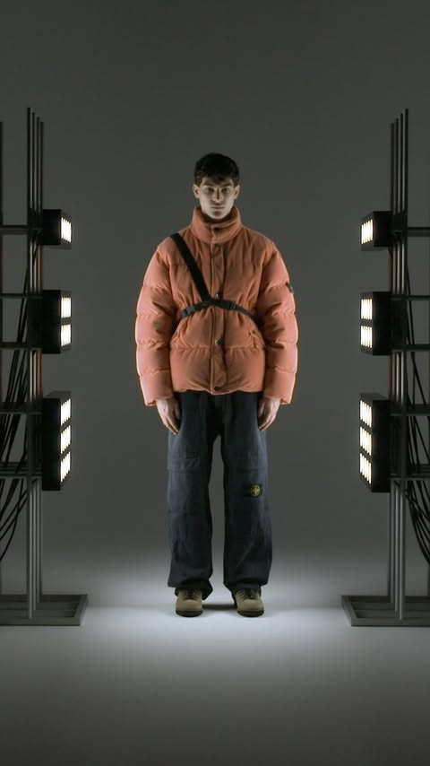 A person wearing a salmon-colored puffer jacket and jeans stands between two metal light stands in a studio environment.