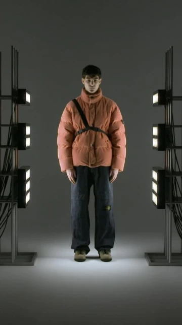 A person wearing a salmon-colored puffer jacket and jeans stands between two metal light stands in a studio environment.