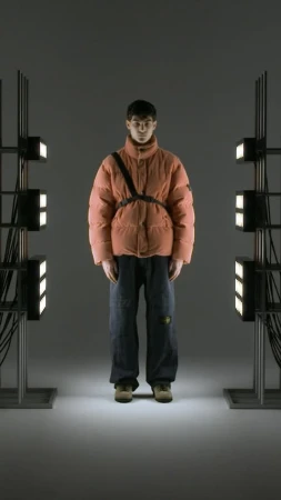 A person wearing a salmon-colored puffer jacket and jeans stands between two metal light stands in a studio environment.