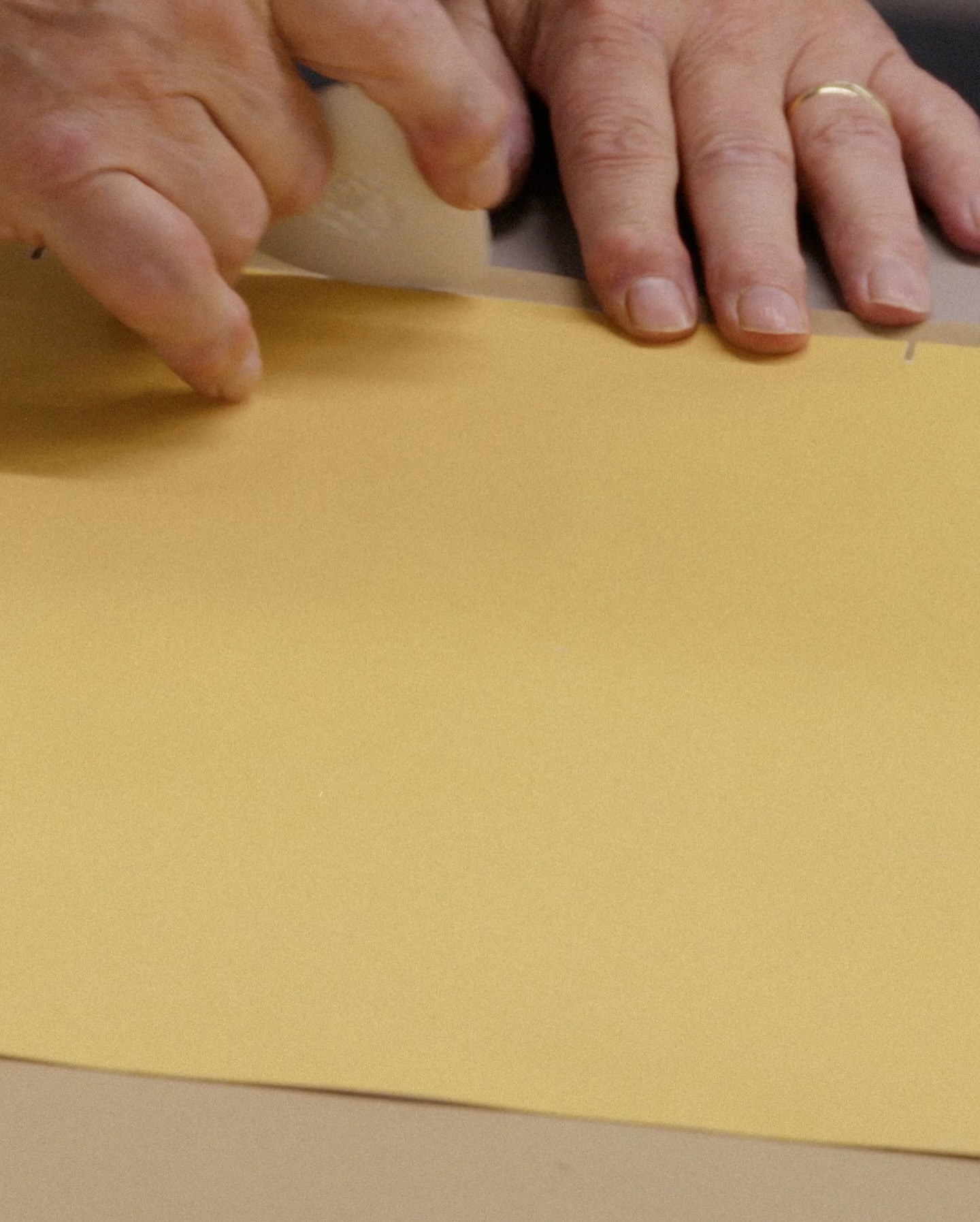 Hands use a craft knife on a sheet of gold paper, cutting carefully on a table surface in an indoor setting for a craft project.