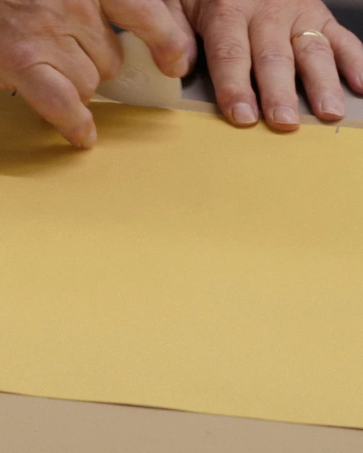Hands use a craft knife on a sheet of gold paper, cutting carefully on a table surface in an indoor setting for a craft project.