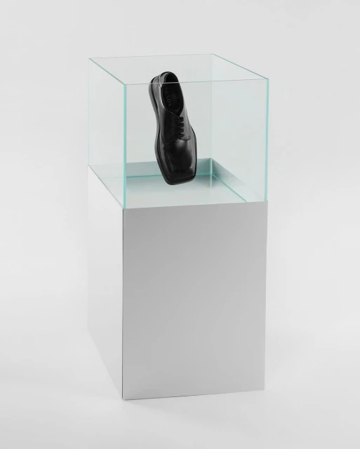 Studio shot of a single black leather dress shoe on display inside a clear glass display case on a white cubical pedestal against white.