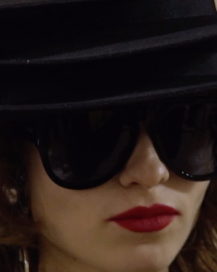 Close-up portrait of a woman wearing black sunglasses, a dark fedora hat, and bright red lipstick.