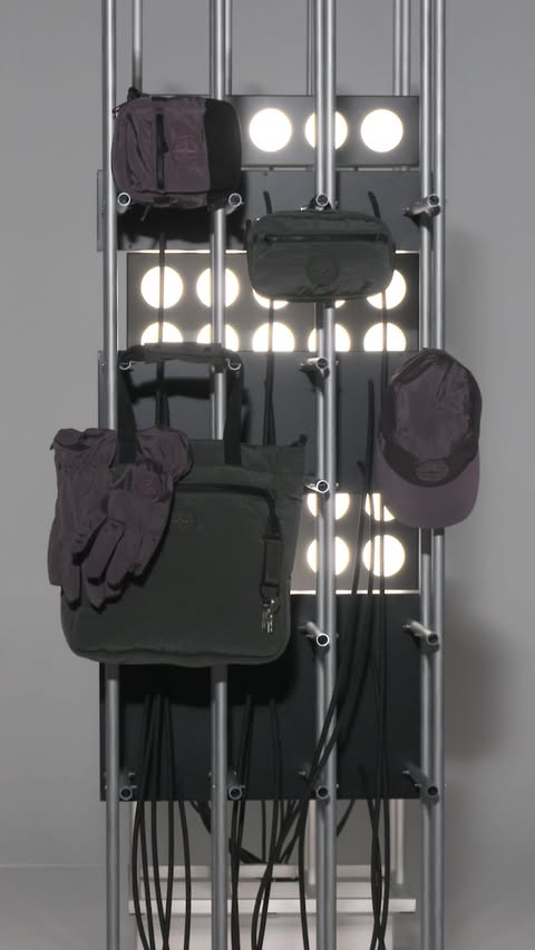 An assortment of bags, gloves, and a cap are arranged on a metal rack against a backdrop of illuminated circles in a studio setting.