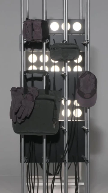 An assortment of bags, gloves, and a cap are arranged on a metal rack against a backdrop of illuminated circles in a studio setting.