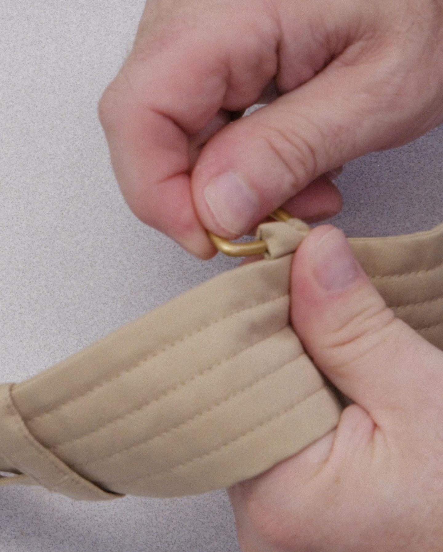 Close-up of hands fastening a tan strap with a gold metal ring on a neutral background, showcasing details and textures of the materials.
