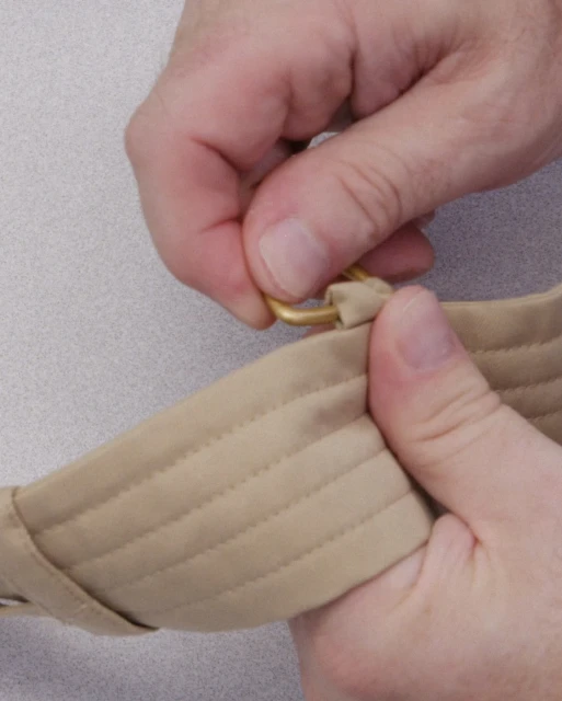 Close-up of hands fastening a tan strap with a gold metal ring on a neutral background, showcasing details and textures of the materials.
