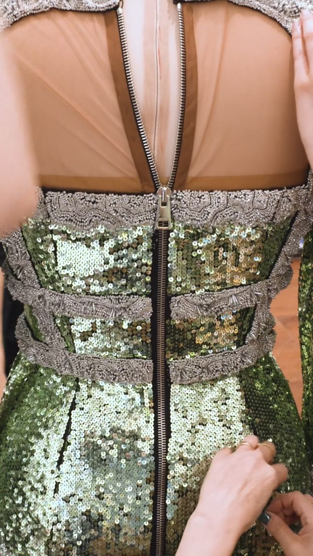 Close-up of a green sequin dress being adjusted, featuring a prominent zipper down the back, lace trim, and sheer fabric panel.