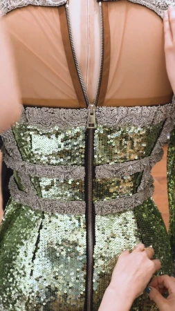 Close-up of a green sequin dress being adjusted, featuring a prominent zipper down the back, lace trim, and sheer fabric panel.
