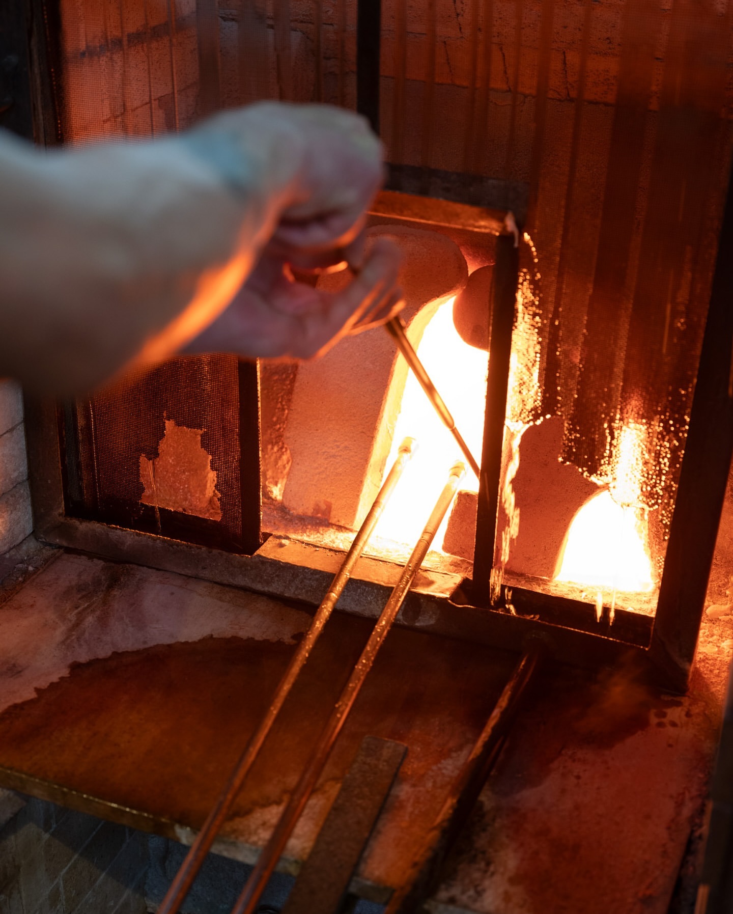 Artisan shaping molten glass with tools in a fiery furnace, creating a handcrafted piece. Intense heat and precision characterize this glassblowing process.