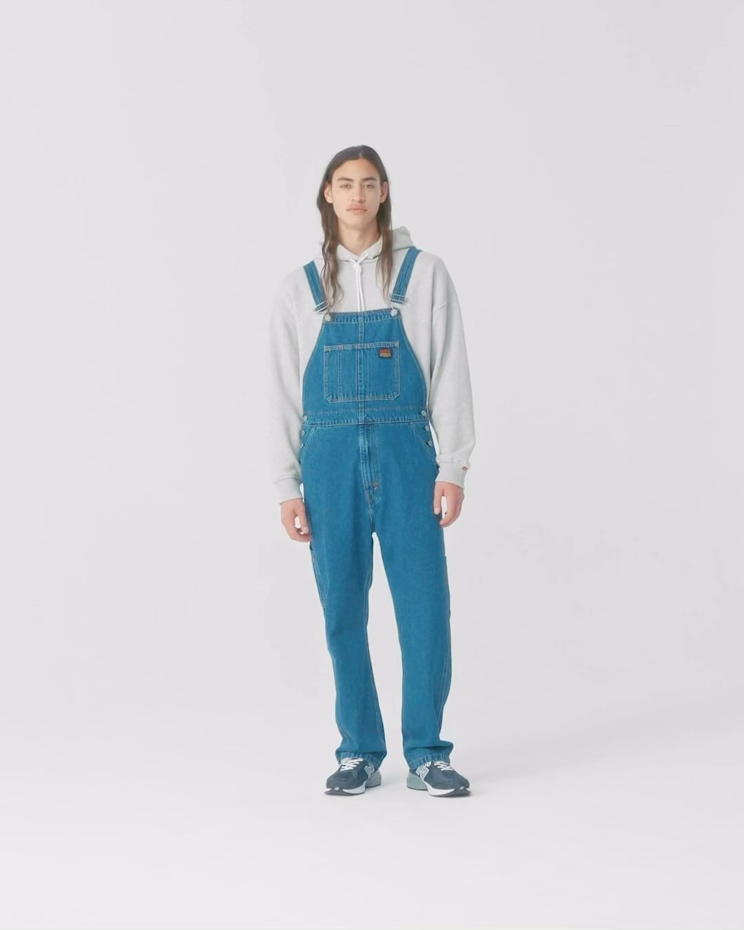 Full-length studio portrait of a man wearing denim overalls and a grey hoodie against a plain white background.