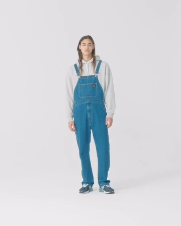 Full-length studio portrait of a man wearing denim overalls and a grey hoodie against a plain white background.