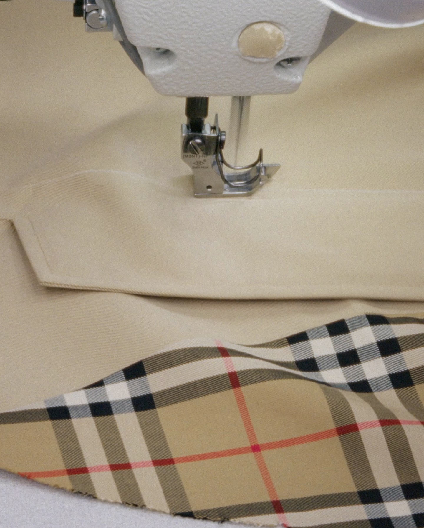 Sewing machine stitching cream-colored fabric next to tan and black tartan fabric. Close-up of fabric and machine.