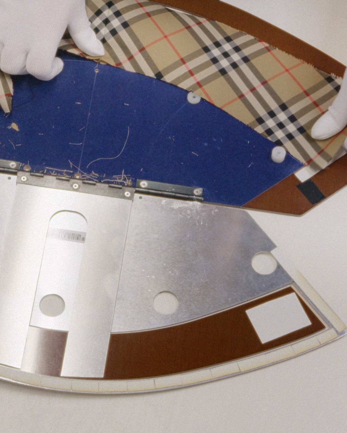 Close-up of hands in gloves adjusting a Burberry pattern fabric with blue backing on a metal cutting machine for garment making.