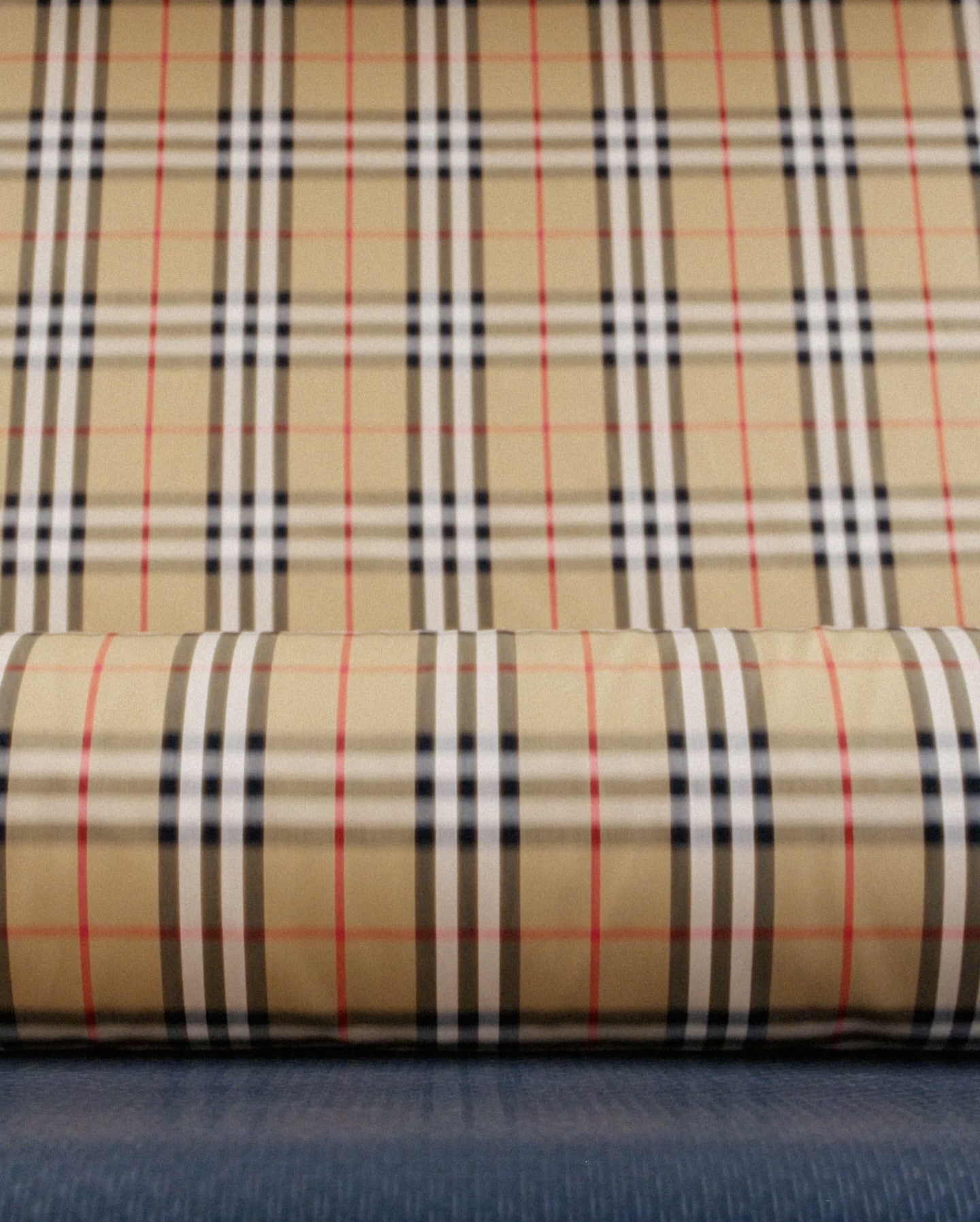 Close-up of a Burberry pattern fabric roll featuring tan, white, black, and red stripes forming a classic plaid design on a textured surface.