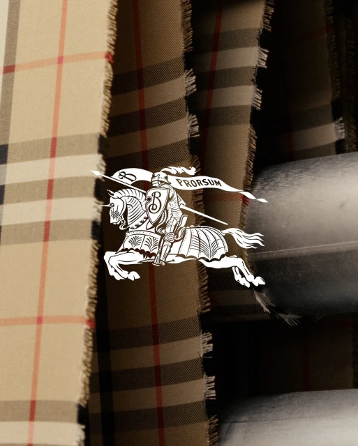 Beige Burberry plaid fabric with blurred metal in the background. Fringed edges and a white knight logo add visual interest.