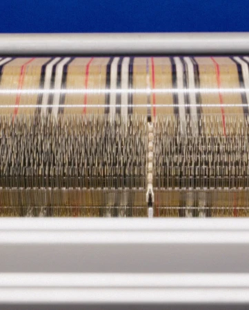 Close-up of weaving machine creating a plaid textile pattern in cream, black, and red, demonstrating textile manufacturing and design.