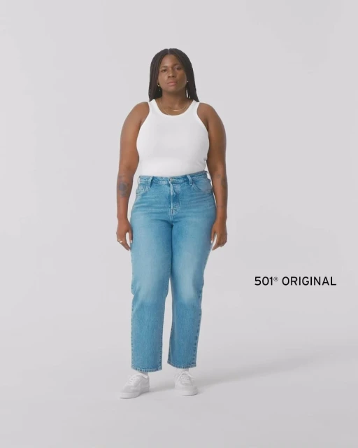 Woman standing in a photo studio wearing a white tank top, jeans, and white shoes, with the text "501 Original" visible.