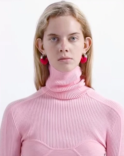 Blonde woman in a pink turtleneck sweater and hoop earrings poses in a close-up studio portrait against a bright white background.