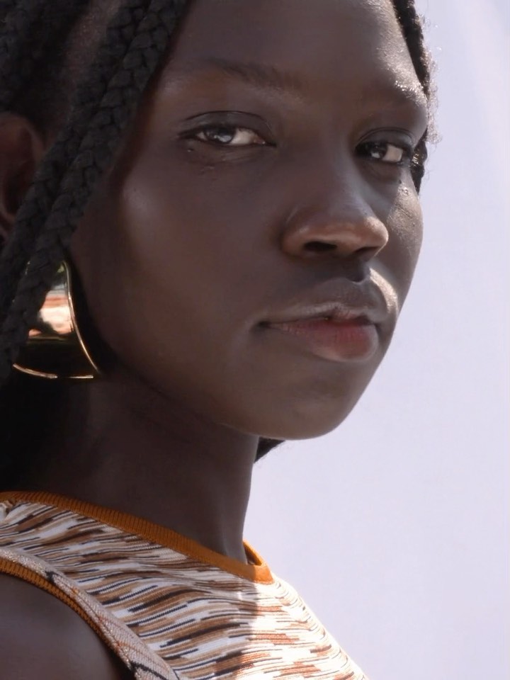 Close-up portrait of a black woman with braided hair wearing a patterned top and large gold hoop earrings against a bright sky.