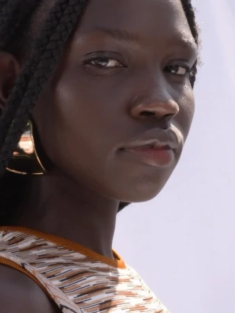 Close-up portrait of a black woman with braided hair wearing a patterned top and large gold hoop earrings against a bright sky.