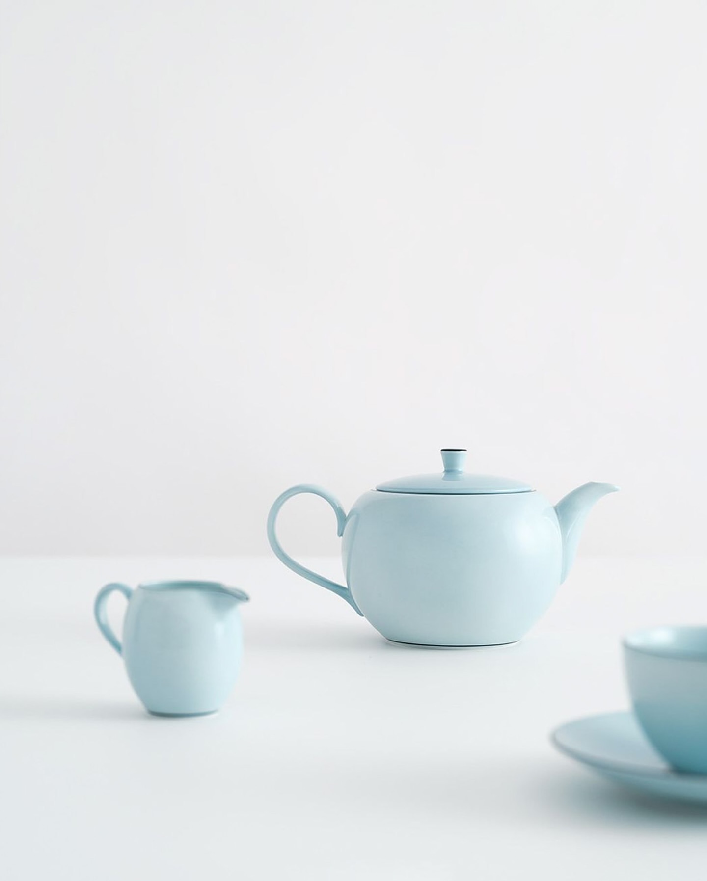 Still life of light blue teapot, creamer, and cup with saucer arranged on white table against plain white background.