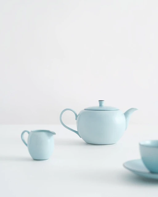 Still life of light blue teapot, creamer, and cup with saucer arranged on white table against plain white background.