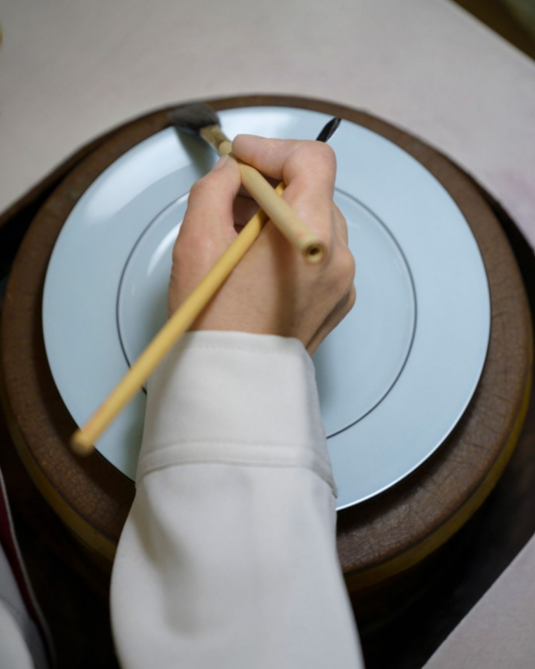 Close-up of an artist's hands carefully painting a delicate line on a light blue plate, using a fine brush and focused technique.