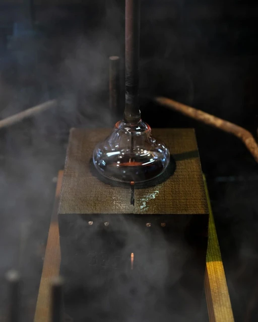 Close up of glassblowing process with molten glass on a metal rod being shaped above a wood surface in an industrial setting with steam.