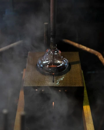 Close up of glassblowing process with molten glass on a metal rod being shaped above a wood surface in an industrial setting with steam.