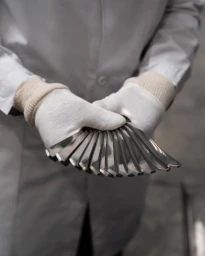 Person wearing a light-coloured coat and white gloves holding a fan of polished metal blades in a workshop setting.