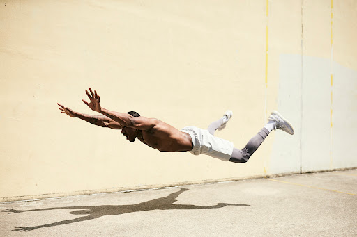 Athletic, shirtless man in mid-air doing a horizontal push-up with arms extended, legs out, on a tan and grey textured surface outside.