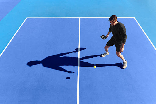 Man playing pickleball on a blue court with white lines; athletic male with paddle, yellow ball, and his shadow visible.
